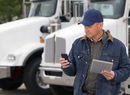 A truck driver wearing a blue jacket and blue cap stands in front of several white trucks, looking at his smart phone.