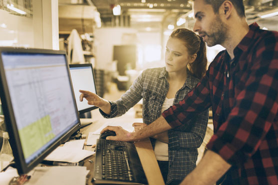 A man and a woman wearing work clothes, are standing in front of a computer, discussing what is on the screen.