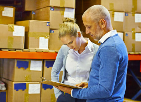 A man and a woman stand in a warehouse, looking down at a clipboard.