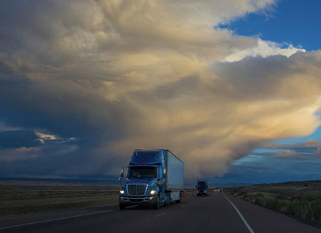 As evening sets, a blue semi-truck with its lights on delivers an expedited freight shipment.