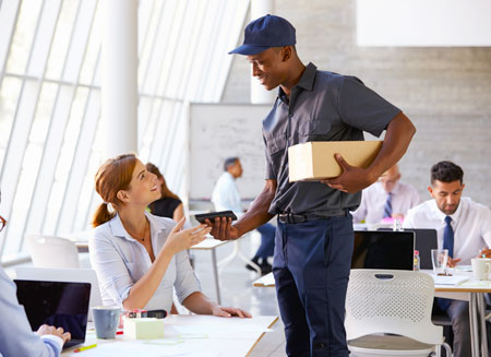 A man wearing a uniform delivers a small package to a woman in an office.